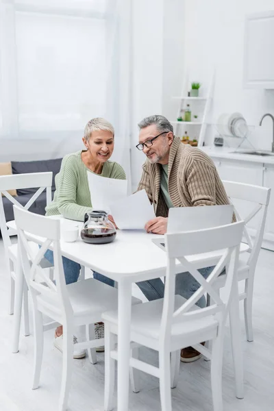 Alegre mujer mayor sosteniendo papel cerca de marido, portátil y café en la cocina - foto de stock