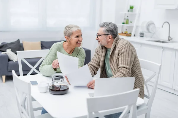 Happy senior woman holding document near husband and devices in kitchen — Stock Photo