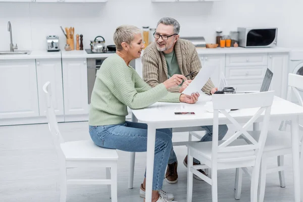 Positive pensioners holding documents near devices in kitchen — Stock Photo