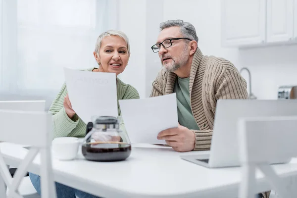 Positive elderly woman holding document near husband, laptop and blurred coffee in kitchen — Stock Photo