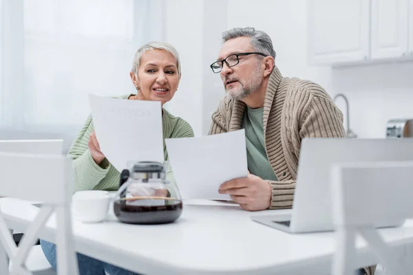 Mujer anciana positiva sosteniendo documento cerca de marido, portátil y café borroso en la cocina - foto de stock