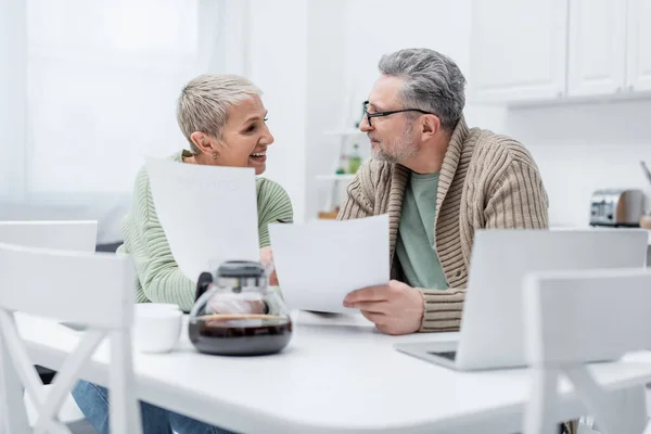 Pensioners holding documents near devices and coffee in kitchen — Stock Photo