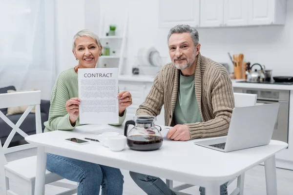 Pensionati in possesso di contratto e guardando la fotocamera vicino a dispositivi e caffè in cucina — Foto stock