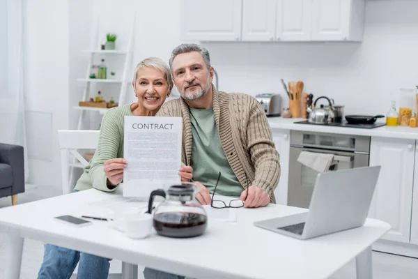 Sonriente mujer mayor celebración de contrato cerca de marido y dispositivos en la cocina - foto de stock