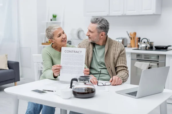 Fröhliche Seniorin mit Vertrag und Blick auf Ehemann in der Nähe von Gadgets und Kaffee in der Küche — Stockfoto