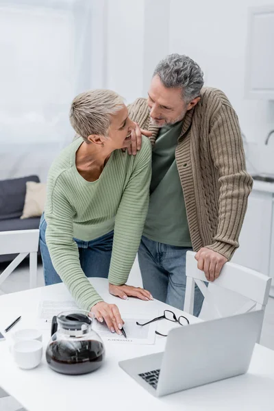 Rentner blickt lächelnde Ehefrau an und zeigt auf Vertrag neben Laptop und Kaffee in Küche — Stockfoto