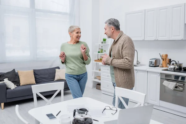 Cheerful senior woman dancing near husband, papers and devices in kitchen — Stock Photo