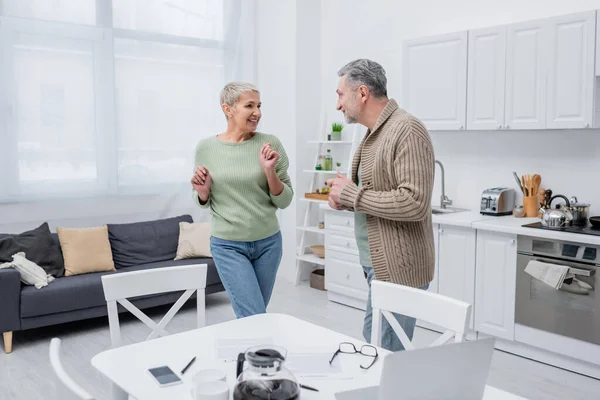 Donna anziana allegra che balla vicino a marito, carte e dispositivi in cucina — Foto stock