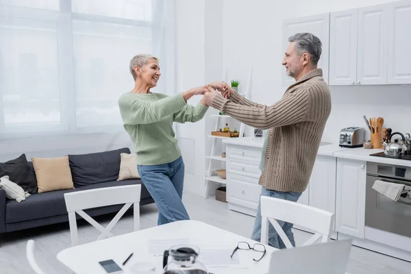 Pareja positiva bailando cerca de papeles y dispositivos en la cocina - foto de stock