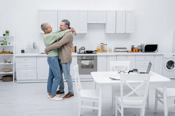 Vista laterale della donna anziana che abbraccia il marito vicino a documenti e dispositivi in cucina — Foto stock