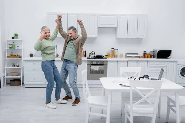 Pareja feliz bailando cerca de papeles y gadgets en la cocina - foto de stock