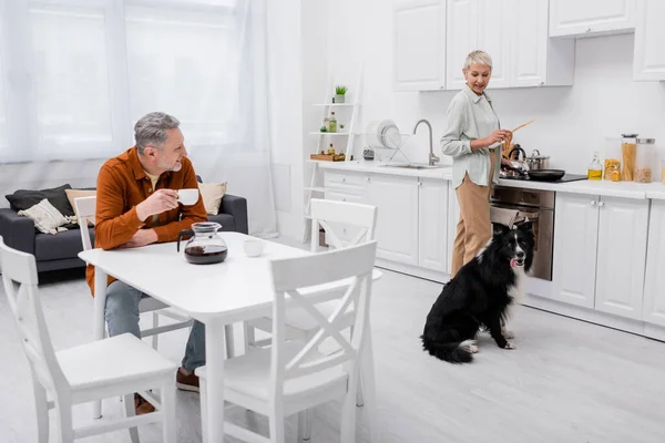 Uomo sorridente che tiene una tazza di caffè vicino alla moglie che cucina e collie di confine in cucina — Foto stock