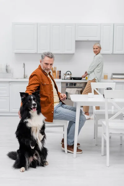 Mature man petting border collie near cup of coffee and blurred wife in kitchen — Stock Photo