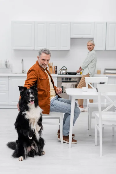 Maduro hombre acariciando frontera collie cerca de taza de café y borrosa esposa en cocina - foto de stock