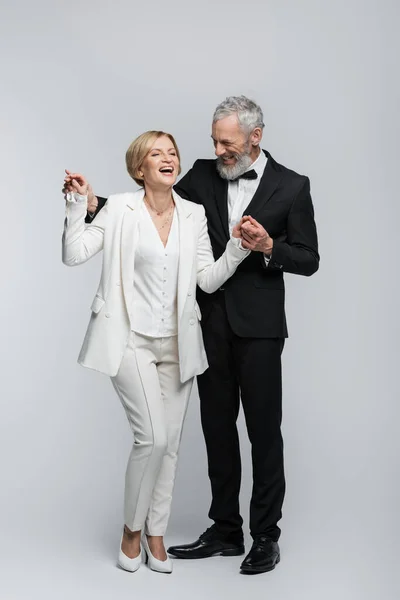 Full length of cheerful groom holding hands of bride in suit on grey background — Stock Photo