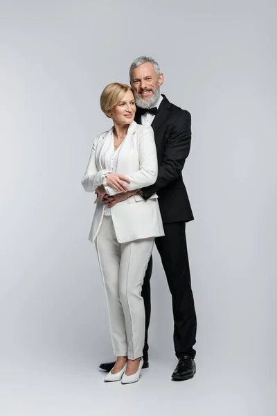 Positive groom hugging mature bride in suit on grey background — Stock Photo