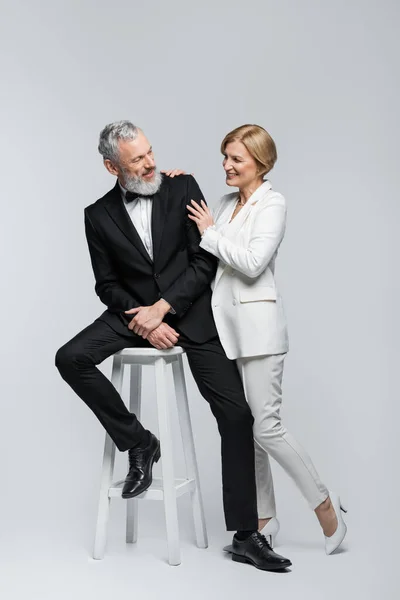 Sonriente novia madura abrazando elegante novio sentado en la silla sobre fondo gris - foto de stock