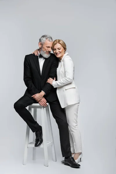 Middle aged bride hugging bearded groom sitting on chair on grey background — Stock Photo