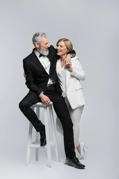 Positive mature groom sitting on chair and hugging bride in suit on grey background — Stock Photo