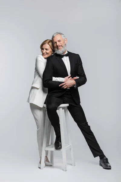 Pleine longueur de mariée mature souriante embrassant marié assis sur la chaise sur fond gris — Photo de stock