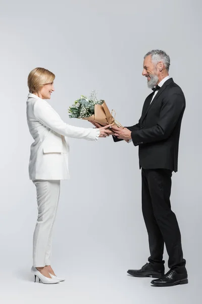Vista lateral del novio maduro dando flores a la novia sobre fondo gris - foto de stock