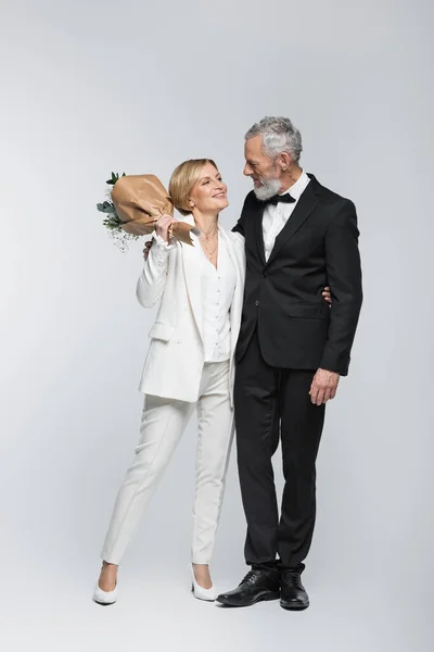 Positivo maturo sposa tenendo bouquet di nozze e abbracciare lo sposo su sfondo grigio — Foto stock
