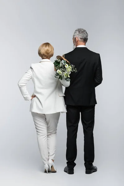 Visão traseira da noiva de terno segurando buquê de casamento perto do noivo no fundo cinza — Fotografia de Stock