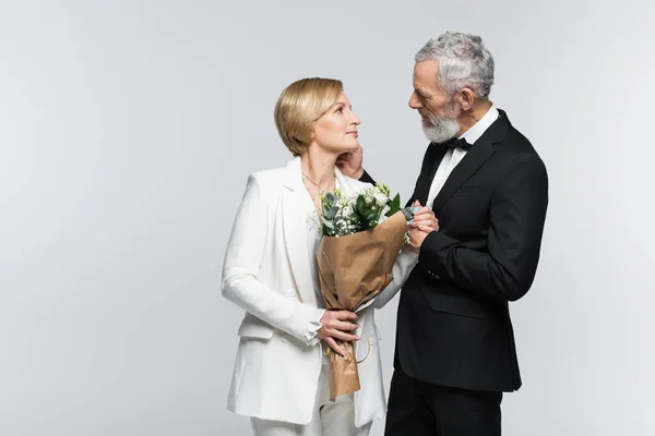 Side view of mature groom touching bride with wedding bouquet isolated on grey — Stock Photo