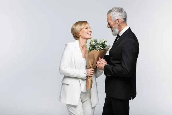 Positive mature bride holding bouquet and hand of groom isolated on grey — Stock Photo