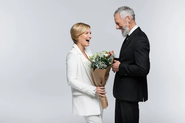 Happy middle aged groom looking at bride with bouquet isolated on grey — Stock Photo