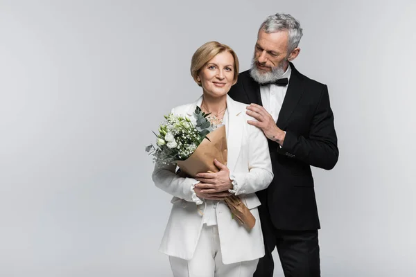 Marié mature en costume étreignant mariée souriante avec bouquet de mariage isolé sur gris — Photo de stock