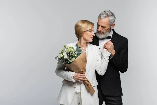 Middle aged groom holding hand of bride with wedding bouquet isolated on grey — Stock Photo