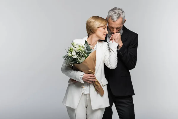 Reifer Bräutigam küsst Hand der Braut im Anzug hält Brautstrauß isoliert auf grau — Stockfoto