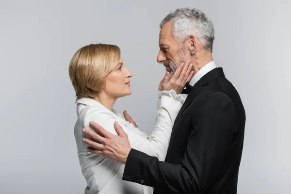 Vue latérale de la mariée mature touchant le visage du marié en costume isolé sur gris — Photo de stock