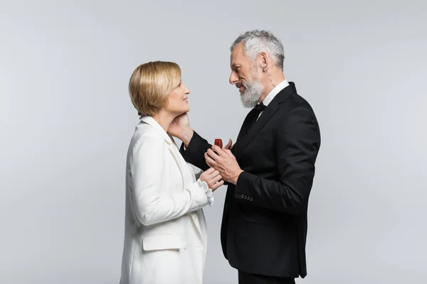 Side view of mature man holding jewelry box and touching smiling woman isolated on grey — Stock Photo