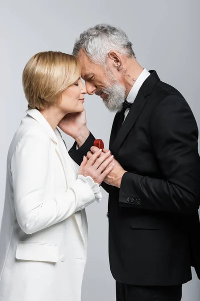 Side view of mature man in suit touching girlfriend and holding jewelry box isolated on grey — Stock Photo