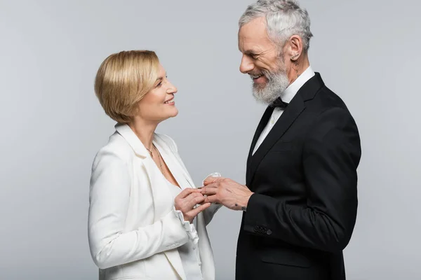 Mariée souriante portant l'anneau de mariage sur le doigt du marié mature isolé sur gris — Photo de stock