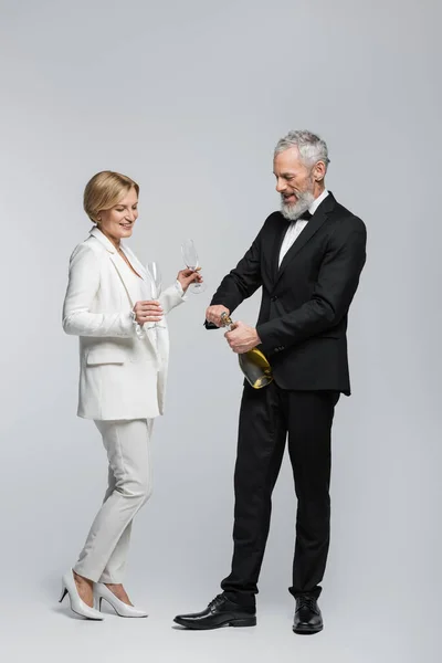 Positive middle aged bride holding glasses near groom with champagne on grey background — Stock Photo