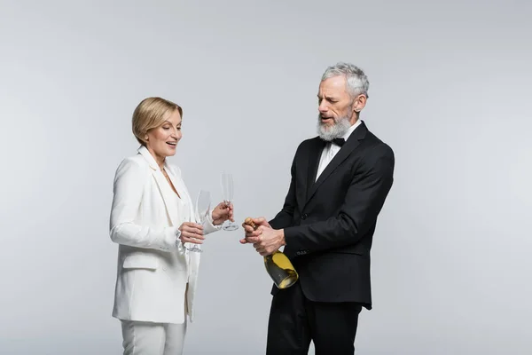 Зрелый жених открывает шампанское рядом с позитивной невестой в стаканах, изолированных на сером — стоковое фото