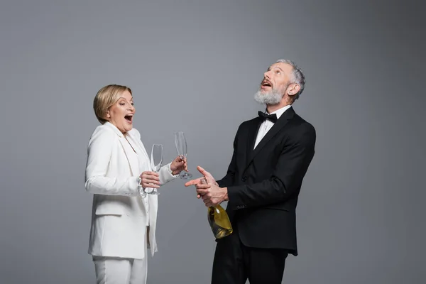 Захоплений зрілий наречений відкриває шампанське біля нареченої в окулярах ізольовані на сірому — стокове фото