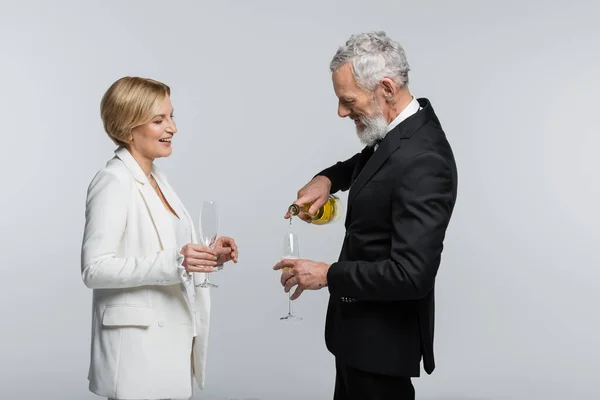 Усміхнений зрілий наречений поливає шампанське біля нареченої склянкою ізольовано на сірому — стокове фото