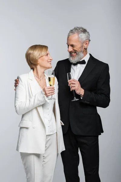 Positive mature groom hugging bride with glass of champagne isolated on grey — Stock Photo