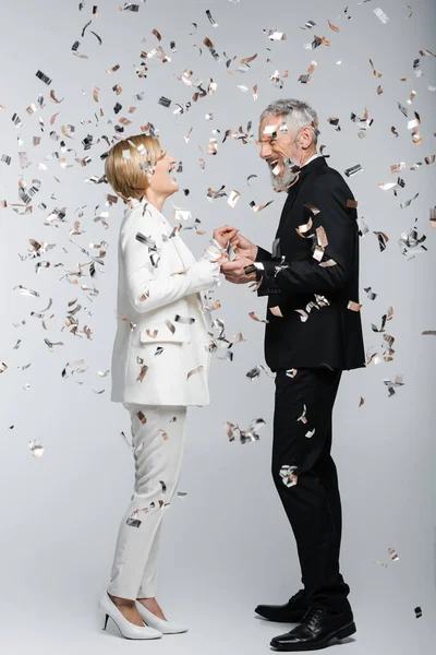 Happy mature groom holding hands of bride under confetti on grey background — Stock Photo