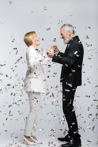Vue latérale du couple mature positif en vêtements de mariage tenant la main sous les confettis sur fond gris — Photo de stock
