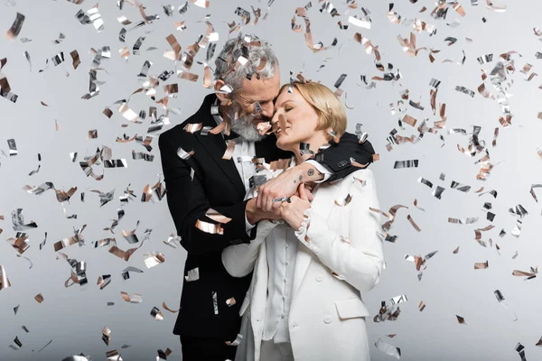 Reifer Bräutigam küsst und umarmt Braut unter Konfetti auf grauem Hintergrund — Stockfoto