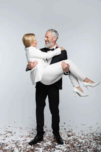 Smiling mature groom holding bride in suit near confetti on grey background — Stock Photo