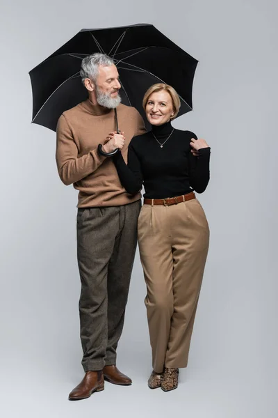 Positive mature woman looking at camera near stylish husband with umbrella on grey background — Stock Photo