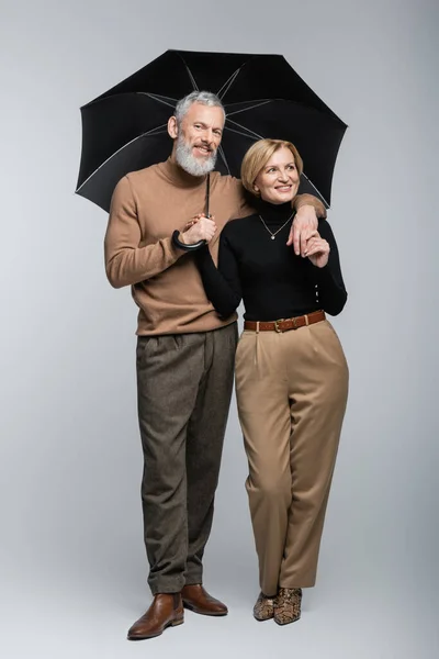 Stylish mature man holding umbrella and hugging cheerful wife on grey background — Stock Photo