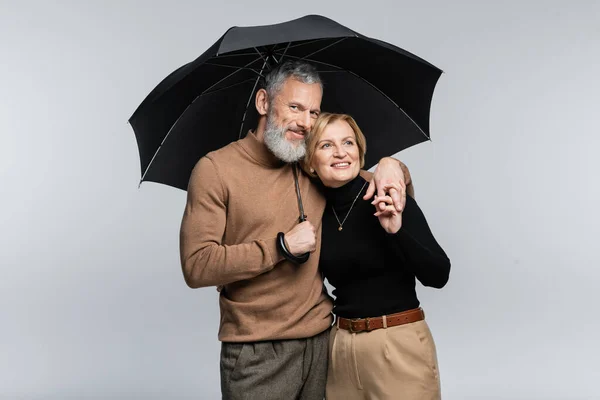 Pareja madura positiva y elegante que sostiene el paraguas y las manos aisladas en gris - foto de stock