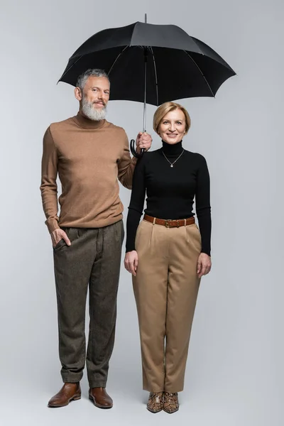 Full length of stylish mature couple with umbrella looking at camera on grey background — Stock Photo